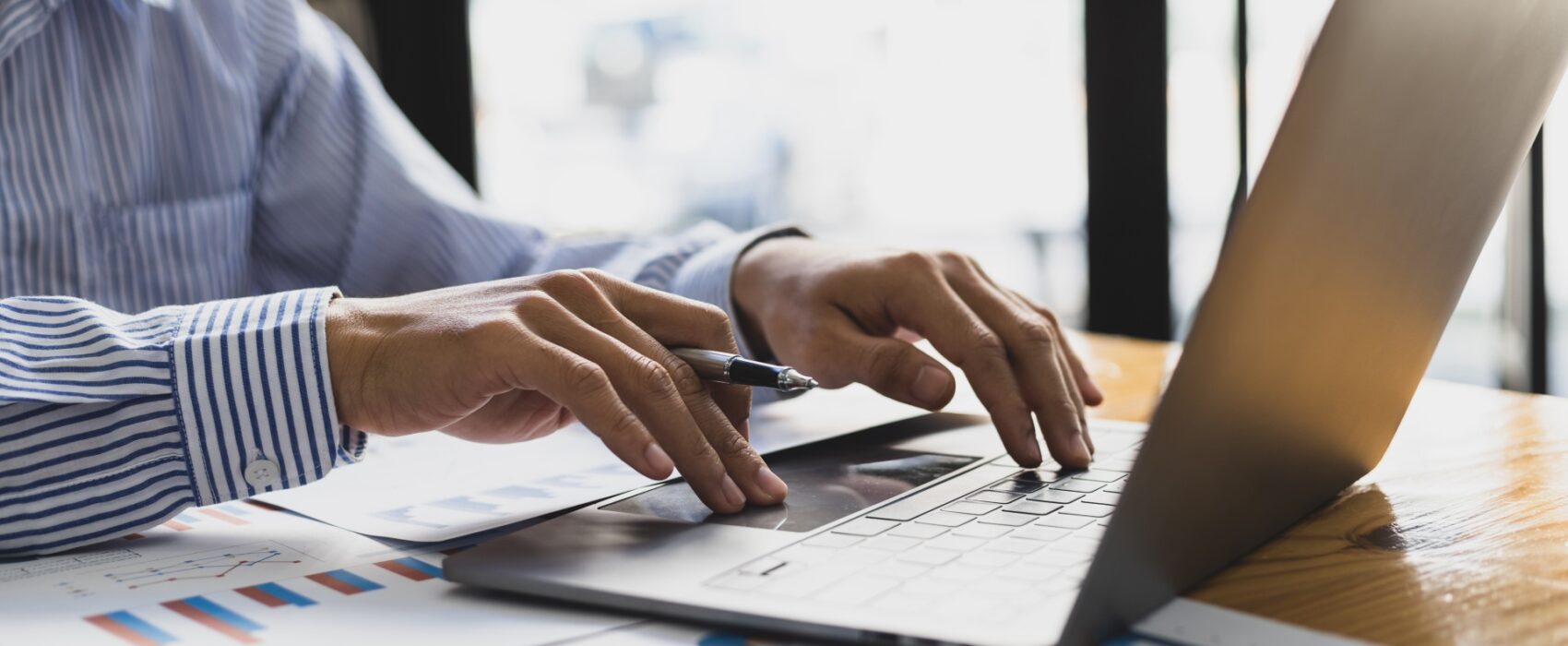 Businesswoman holding a pen and analyzing documents while working on a laptop at her desk. Translation of financial documents.