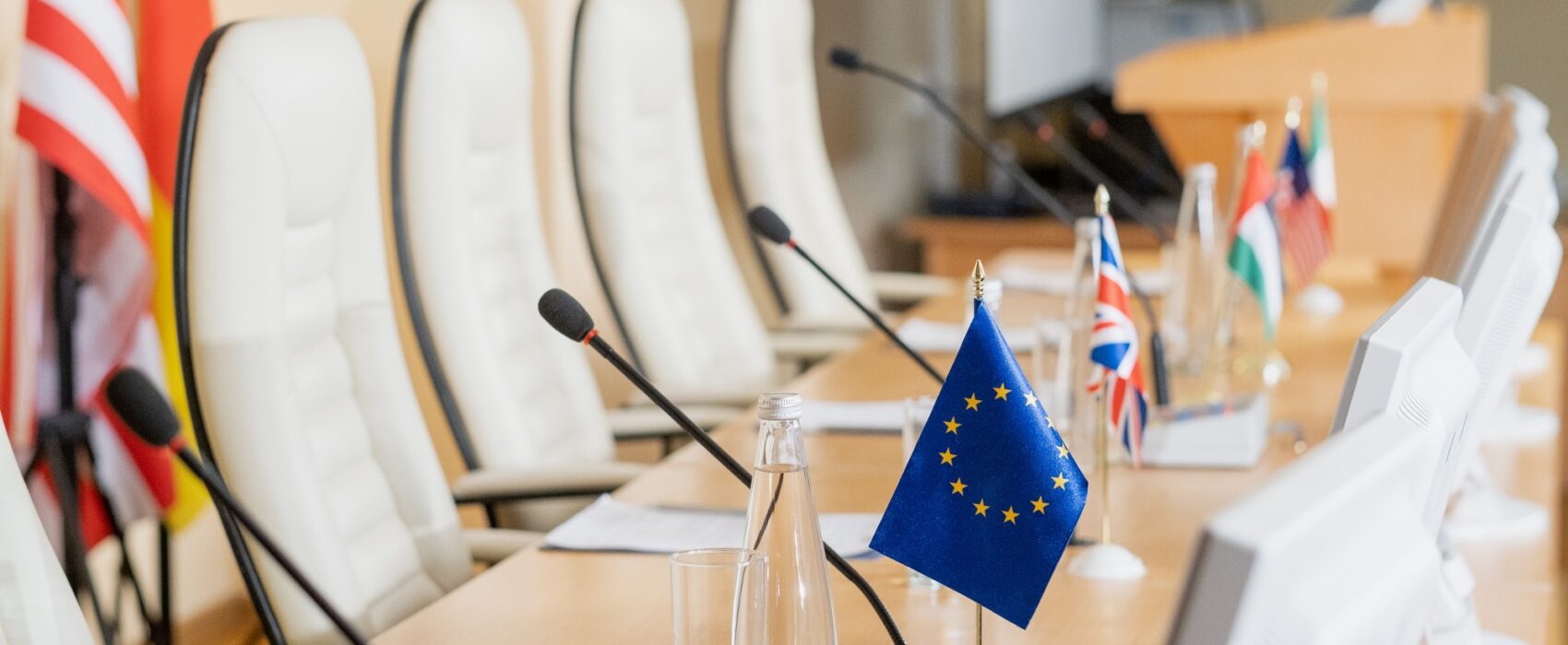 Row of microphones, flags, papers and other on long table in conference hall